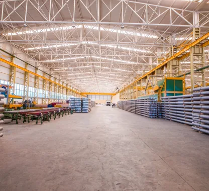 Interior de um amplo galpão fabril com estrutura metálica aparente no teto, exibindo linhas de produção organizadas e estoques laterais de perfis cilíndricos, ilustrando o impacto do layout na ventilação industrial para galpão.
