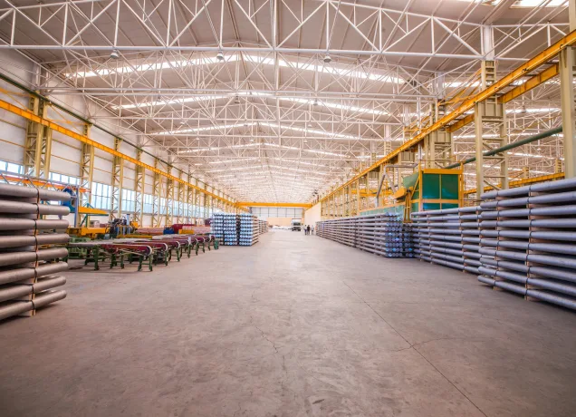 Interior de um amplo galpão fabril com estrutura metálica aparente no teto, exibindo linhas de produção organizadas e estoques laterais de perfis cilíndricos, ilustrando o impacto do layout na ventilação industrial para galpão.